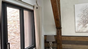 Un dormitorio con ventana, estructura de vigas de madera, una mesita de noche con lámpara y una obra de arte en la pared.
