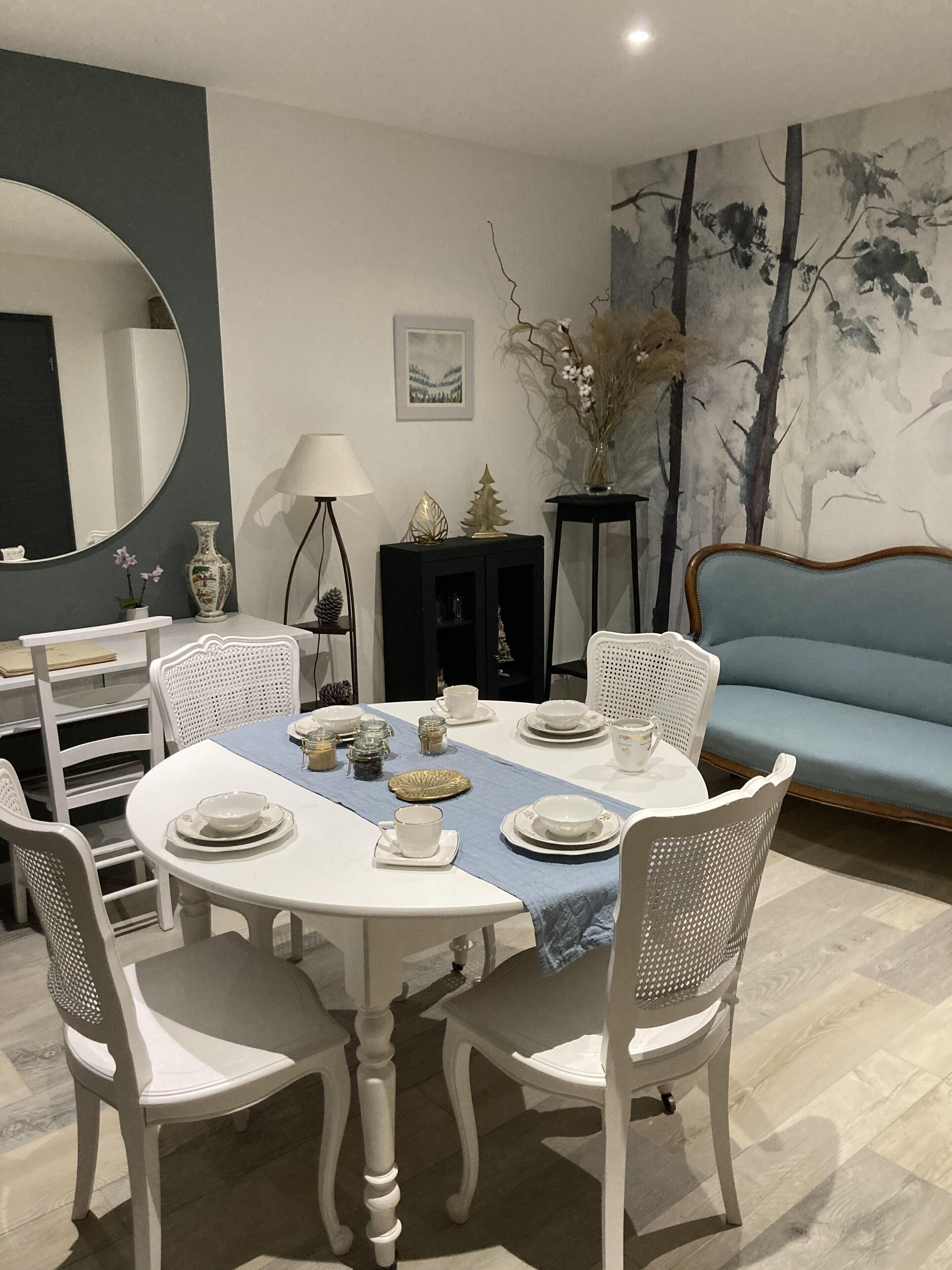 Un comedor con una mesa redonda dispuesta para cuatro personas, rodeada de sillas blancas. Hay un camino de mesa azul, un jarrón con flores secas y un cuadro en la pared.