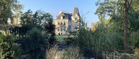 A large, historic building with a prominent tower, surrounded by lush greenery and a small pond.