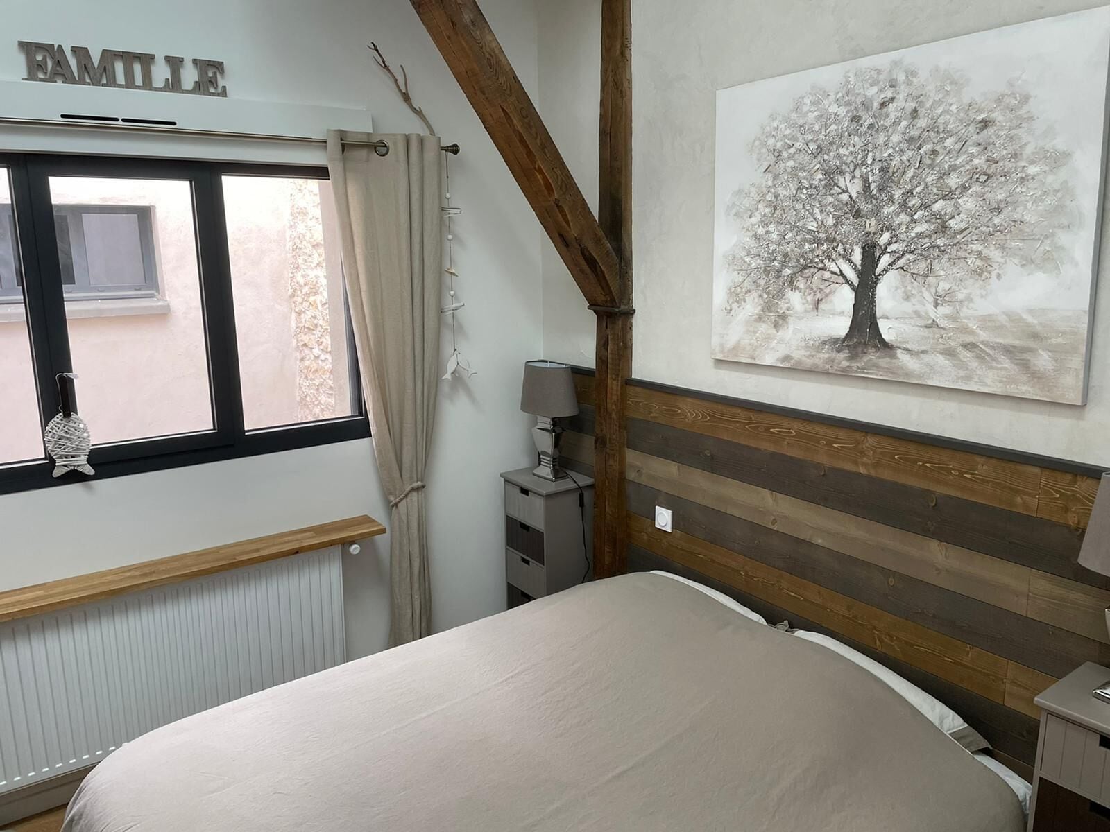 Une chambre à coucher avec un lit, une fenêtre avec des rideaux, un radiateur, une table de chevet avec une lampe et un tableau représentant un arbre, accroché au mur.