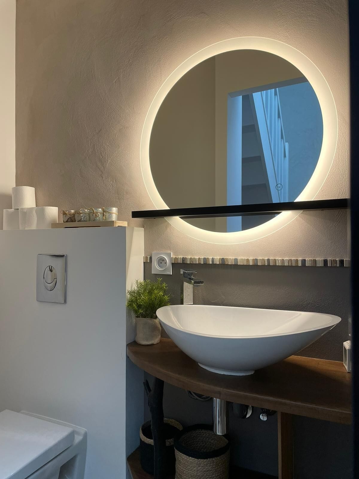 Une salle de bain moderne avec un miroir rond, un lavabo blanc et une petite plante en pot.