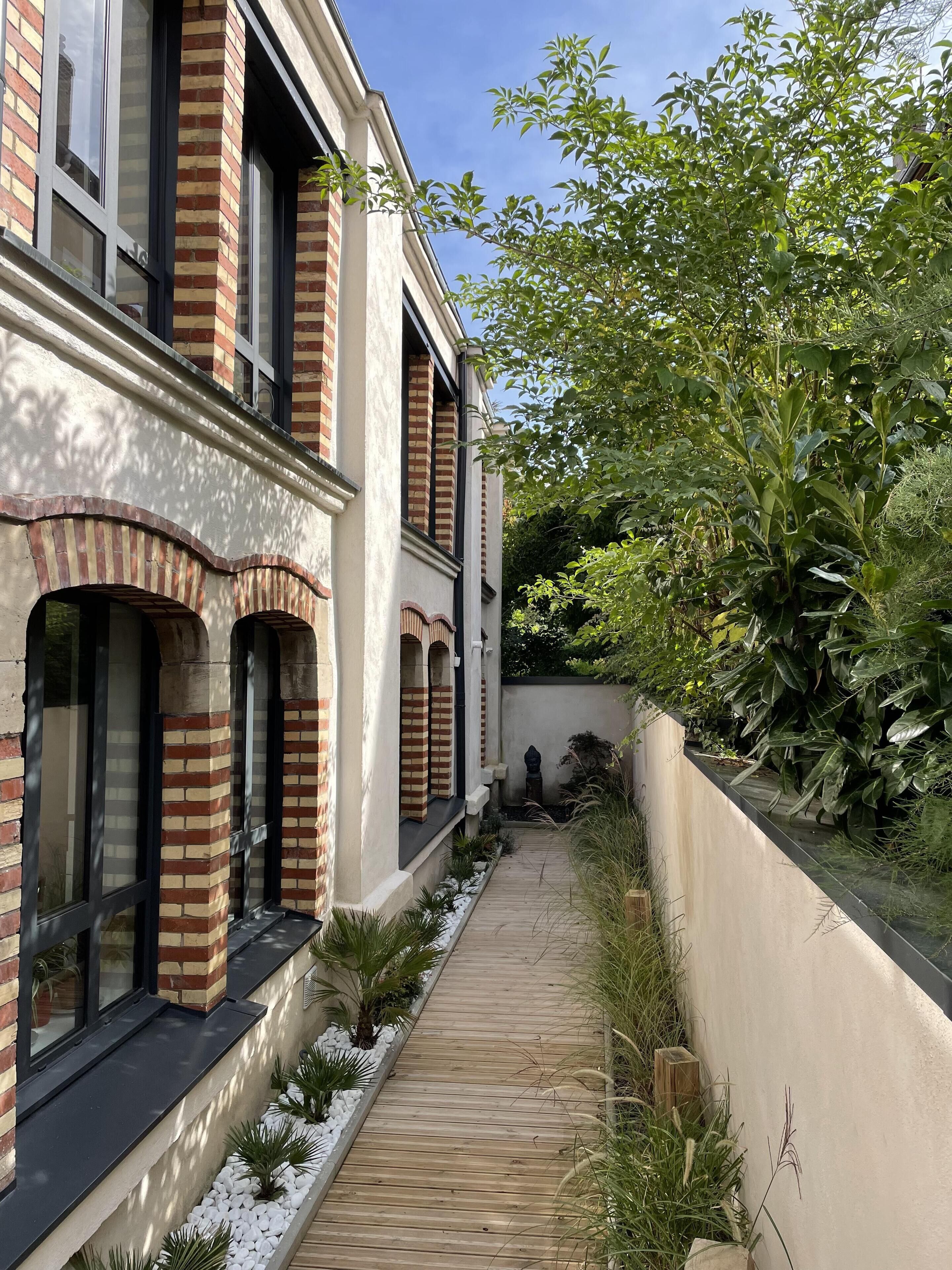 Un edificio moderno con paredes de ladrillo y blanco, amplios ventanales y un sendero de madera que conduce a un jardín con diversas plantas y una pequeña estatua.