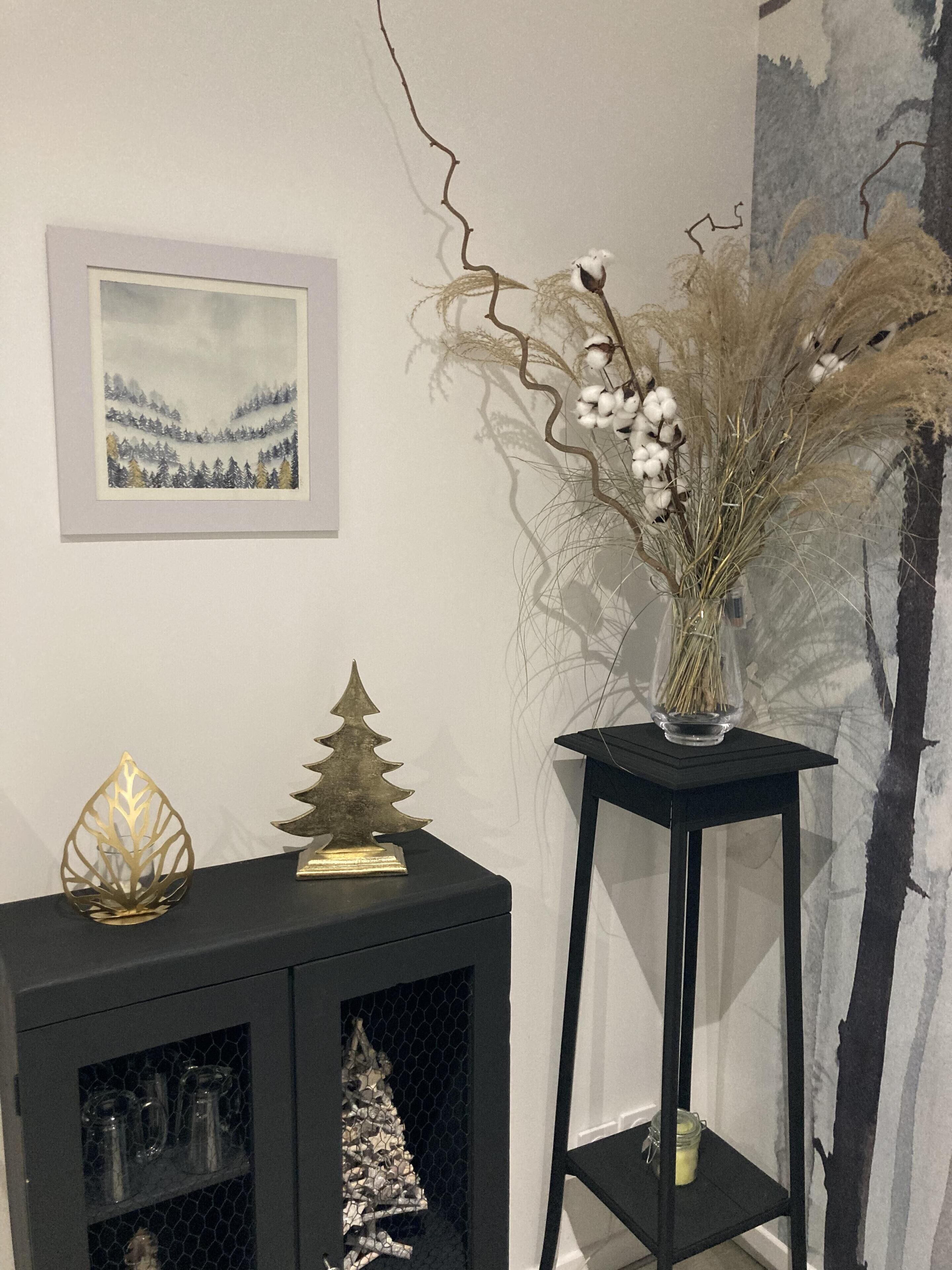 Una mesita auxiliar negra con un árbol decorativo dorado y un florero de vidrio con plantas secas.