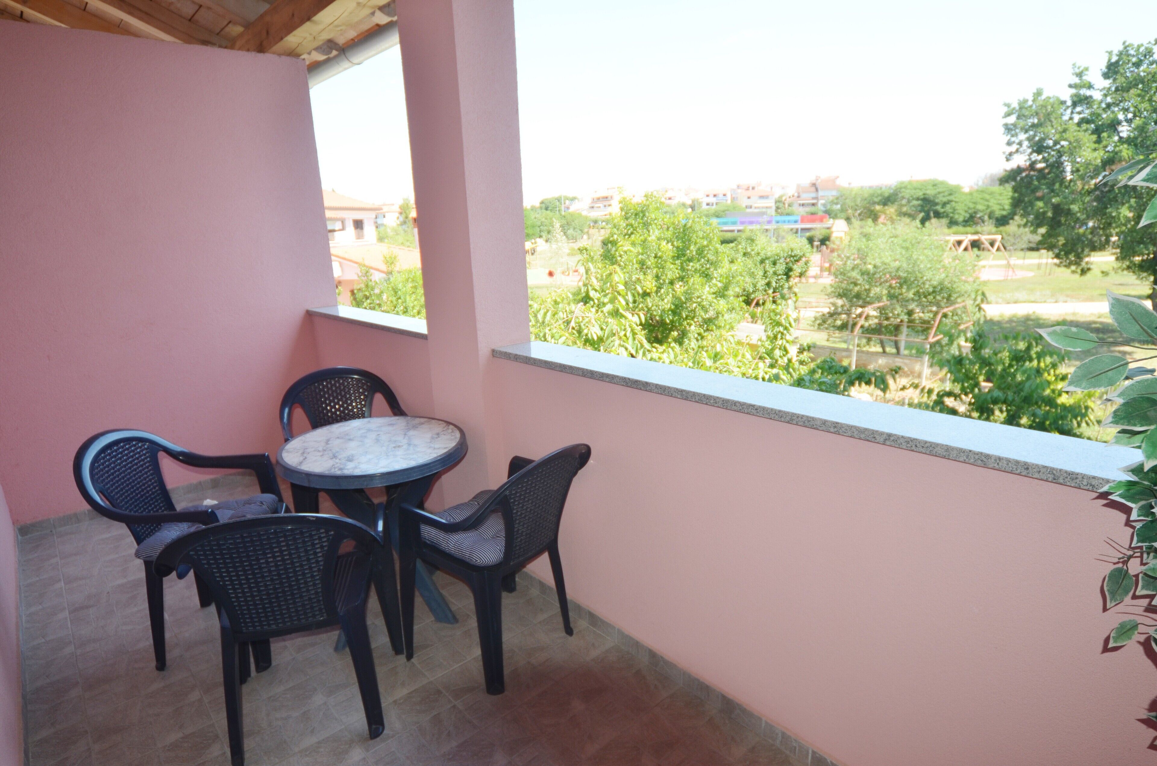 Un balcón con mesa y sillas, una pared rosa y vistas a zonas verdes y edificios.