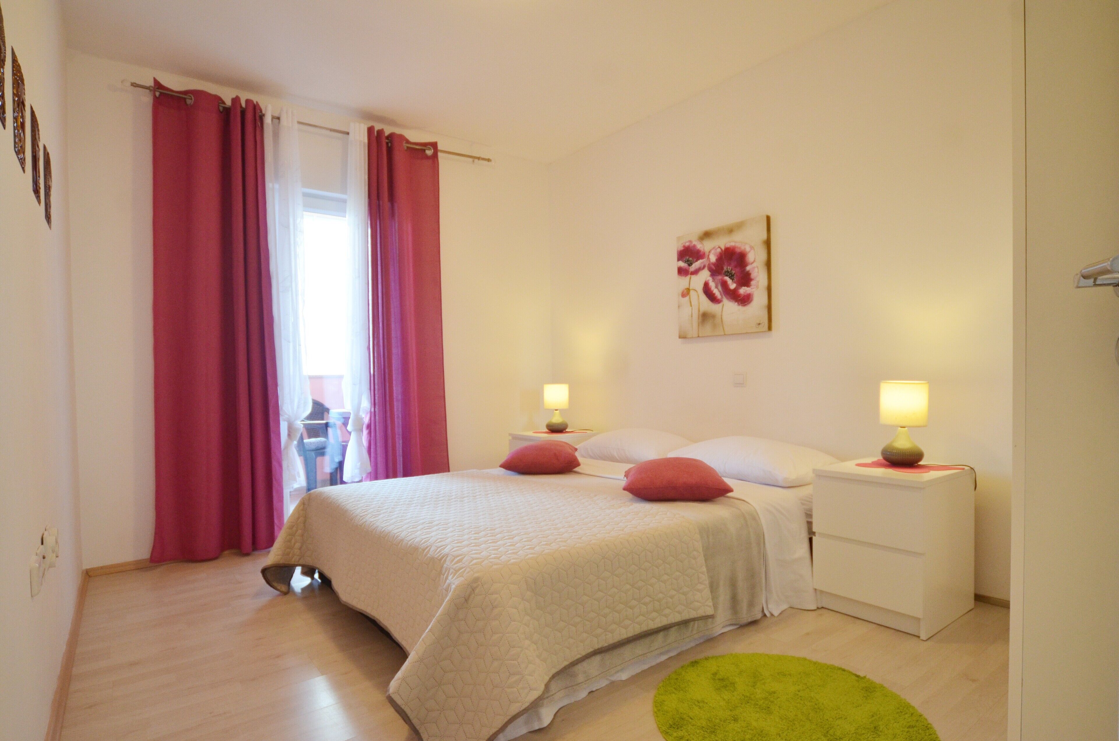 A bedroom with a bed, two bedside tables with lamps, a green rug, and a window with curtains.