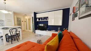 A modern living room with an orange sofa, a dining area with a white table and chairs, and a kitchen with white cabinets and a black TV. - Bea & Leo Apartment in center (Rovinj)