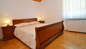 Een slaapkamer met een houten bed, twee nachtkastjes met lampen, een radiator en een raam met dunne gordijnen.