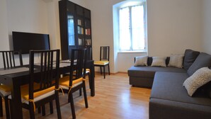 Ein Wohnzimmer mit einem dunkelgrauen Sofa, einem Flachbildfernseher, einem Esstisch mit Stühlen und einem Bücherregal.