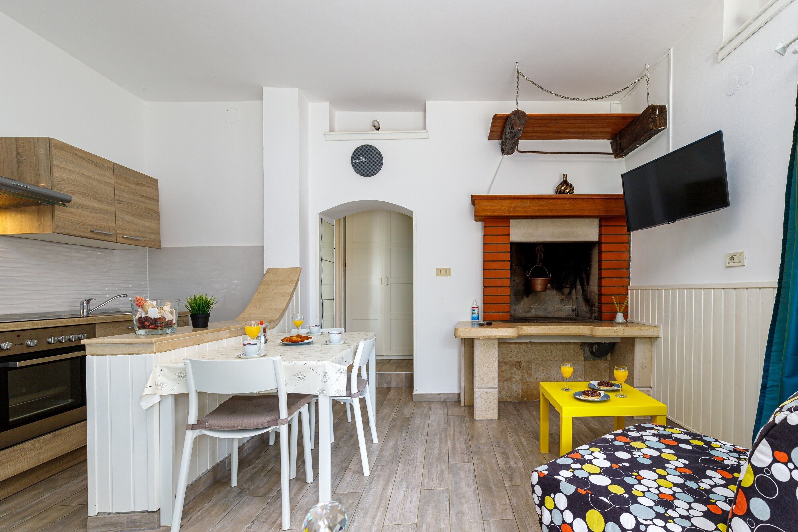 Cocina compacta con comedor, televisor montado en la pared y un pequeño rincón de descanso con un cojín estampado.