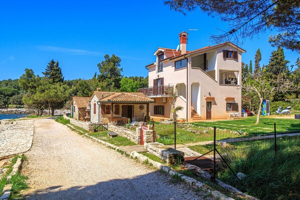 Une maison à deux étages, avec un chemin de pierres menant à l’entrée, entourée de verdure et sous un ciel bleu dégagé.