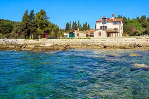 Un bâtiment situé sur la côte, doté d’un mur en pierre, avec une eau d’un bleu limpide et des arbres en arrière-plan.