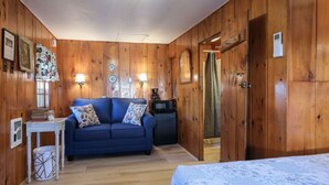 Ein gemütliches Zimmer mit Holzwänden, einem blauen Sofa mit gemusterten Kissen, einem kleinen Tisch mit Büchern und einer Glaseinlage sowie einem Bett mit blauen und weißen Kissen.