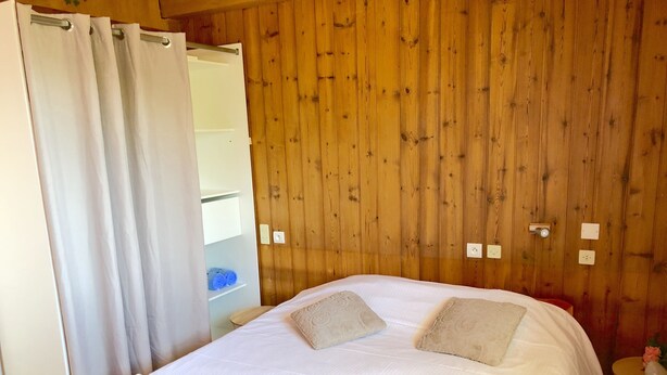 Ein Schlafzimmer mit Holzwand, einem Bett mit weißer Bettwäsche, zwei Kissen, einem Nachttisch mit einer Vase und einem teilweise geöffneten Vorhang.