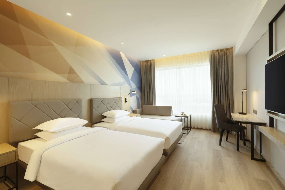 A hotel room with two beds, a desk, a chair, a TV, and a window with curtains.