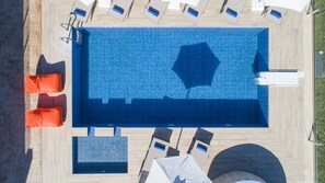 Eine Vogelperspektive auf ein rechteckiges Schwimmbecken mit einem geometrischen Fliesenmuster, umgeben von Liegestühlen und einer Sonnenliege.