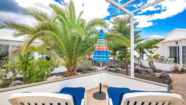 Uma área de pátio com duas cadeiras de lounge brancas, um guarda-sol colorido e uma palmeira.