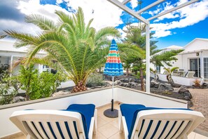 A patio area with two white lounge chairs, a colorful umbrella, and a palm tree. - A3, in a small complex with communal pool 100m from the sea (Charco del Palo)