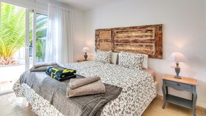 Quarto com uma cama grande, cabeceira de madeira, duas mesas de cabeceira com abajures e vista para palmeiras através de uma porta de correr.
