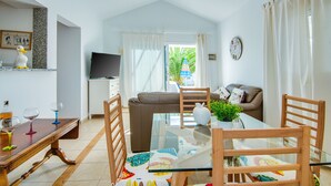Uma sala de estar moderna com uma mesa de jantar de vidro, cadeiras de madeira, um sofá marrom e uma televisão sobre um armário branco.
