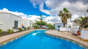 Piscina com bordas azulejadas, rodeada por palmeiras e cadeiras de praia, com edifícios brancos ao fundo.