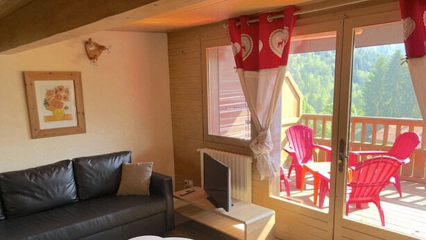 Ein gemütliches Wohnzimmer mit einer schwarzen Ledersofa, einem weißen Couchtisch, einem Fernseher auf einem Schrank und einer Glastür, die auf eine Terrasse mit roten Stühlen führt.