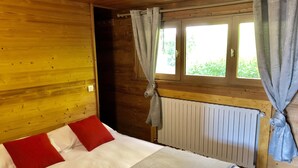 Ein Zimmer einer Holzhütte mit einem Bett, roten und weißen Kissen, einem Fenster mit Vorhängen und einer Heizung.