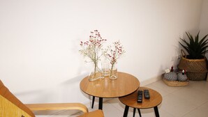 Een kleine, gezellige kamer met een houten stoel, een ronde houten tafel met twee vazen vol bloemen, en een afstandsbediening op de tafel.