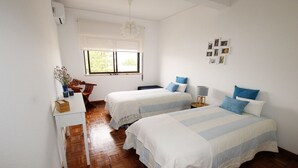 Een slaapkamer met twee bedden, houten vloeren, een raam met zonwering, een tafeltje met een lamp en een muur met foto's.