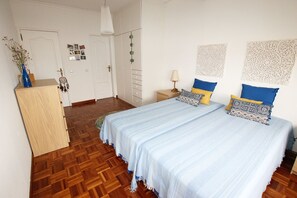A bedroom with a wooden floor, a bed with blue and patterned pillows, a bedside table with a lamp, and a wardrobe.
