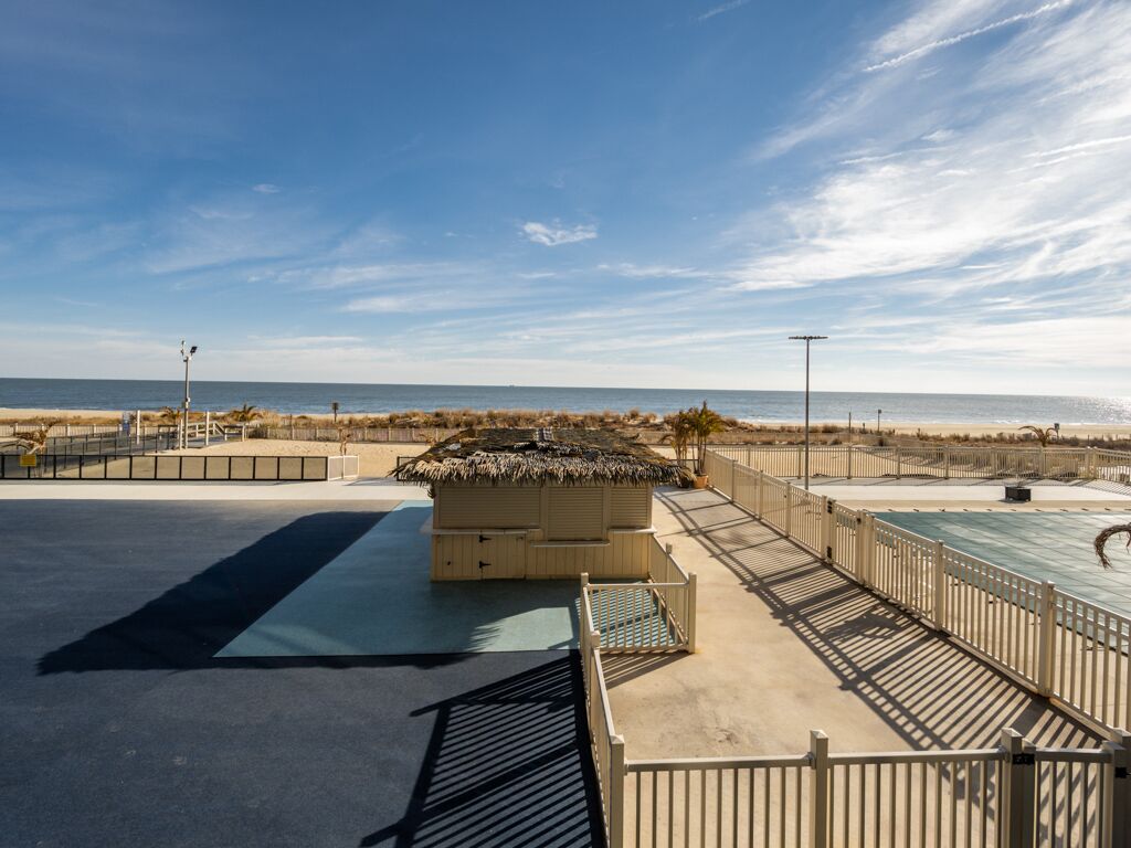 Un espace piscine situé au bord de la plage, doté d’une structure à toit de chaume, d’une piscine et d’une allée piétonne clôturée.