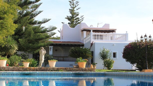 A poolside view of a white building with a tiled roof, surrounded by potted plants and trees.