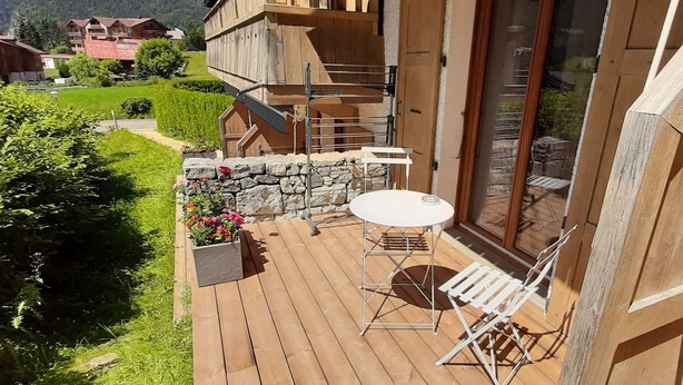 Una terraza de madera con una mesa y sillas, un muro de piedra y vistas a un pueblo de montaña.
