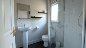 A bathroom with a white sink, toilet, and shower. There is a window with blinds, a mirror above the sink, and a shelf with toiletries. - Drummond (Aberfeldy)