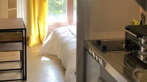 Een moderne kamer met een bed, een televisie, een kitchenette en een groot raam met gordijnen.
