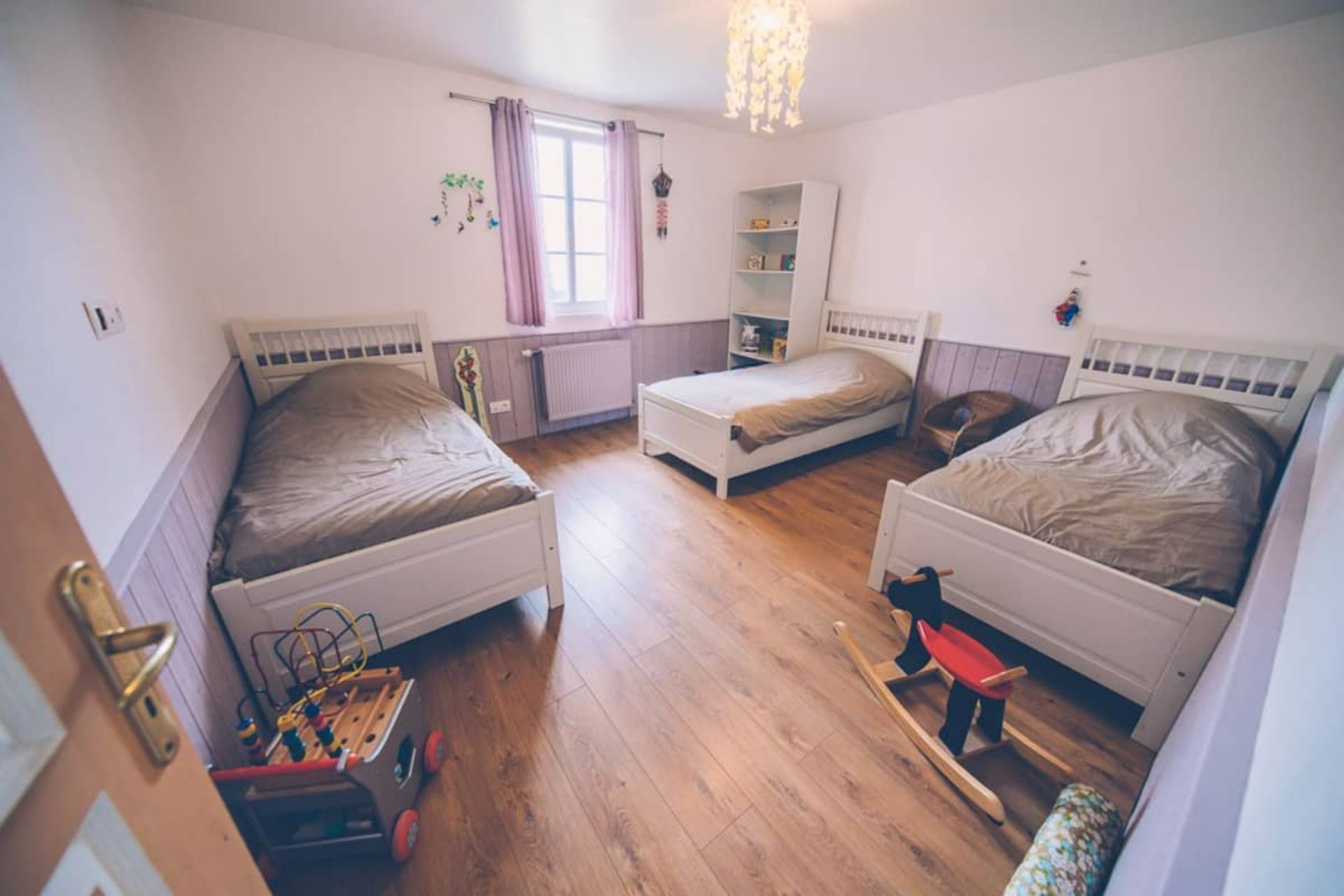 A room with three single beds, a radiator, a window with curtains, and a wooden floor.