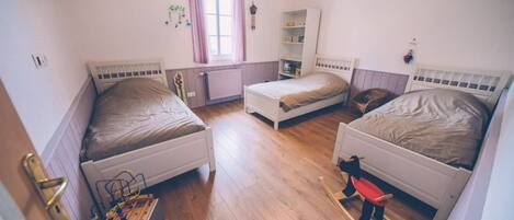 A room with three single beds, a radiator, a window with curtains, and a wooden floor.