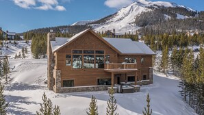 A wooden cabin with a balcony, surrounded by snow and trees, with a mountain in the background.