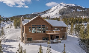 A wooden cabin with a balcony, surrounded by snow and trees, with a mountain in the background.