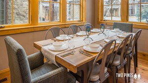 Un comedor con mesa de madera dispuesta para cuatro personas, rodeada de sillas, ventanas amplias con paredes de piedra y vistas al exterior.