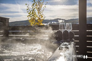 A hot tub with two glasses, surrounded by steam, with a view of mountains and a tree with yellow leaves in the background.