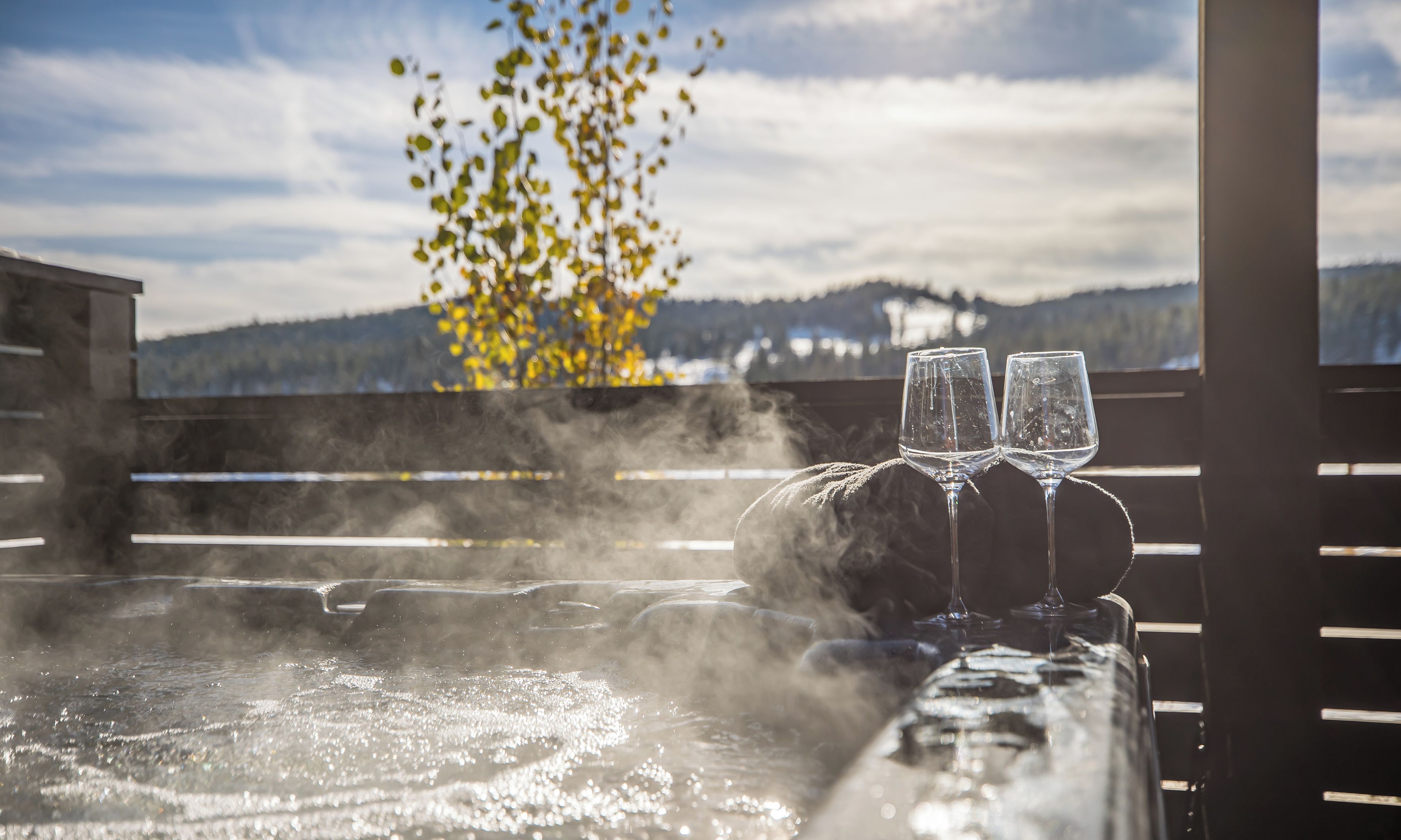 Un bain à remous avec deux verres, entouré de vapeur, offrant une vue sur les montagnes et un arbre aux feuilles jaunes en arrière-plan.