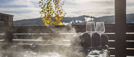 Un bain à remous avec deux verres, entouré de vapeur, offrant une vue sur les montagnes et un arbre aux feuilles jaunes en arrière-plan.