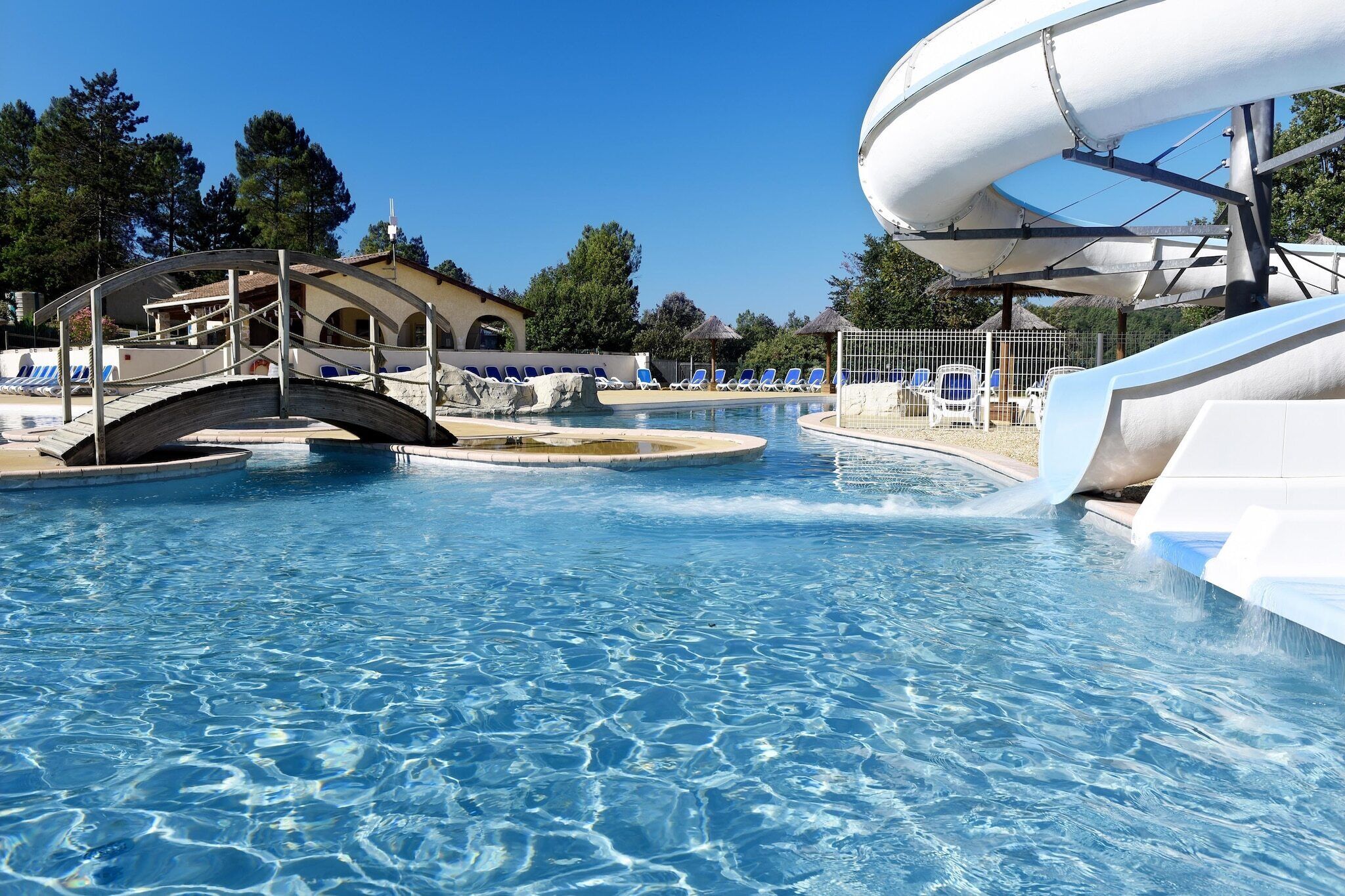 A swimming pool with a water slide, lounge chairs, and a bridge.