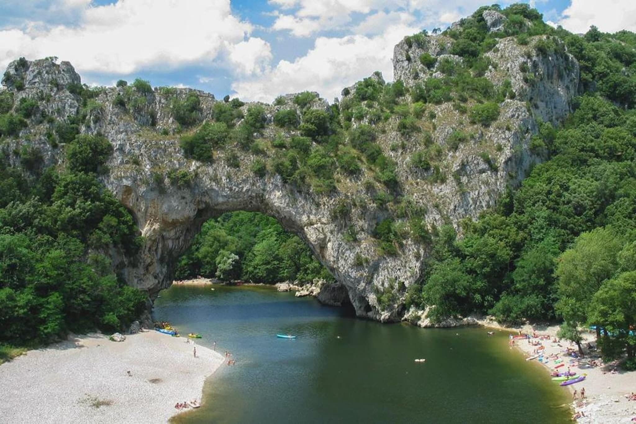 A natural rock arch formation over a body of water with a sandy beach and greenery on the sides.