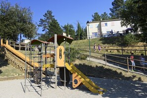 A playground with a slide and climbing structures, surrounded by a fence and trees.