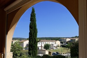 A view through an archway of a residential area with houses, a tall tree, and a clear blue sky.