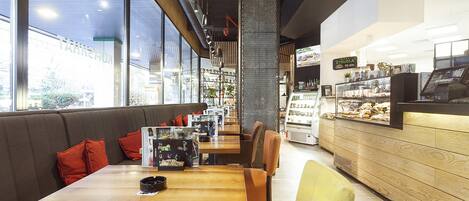 A modern cafe with wooden tables and chairs, a counter with a display case, and a large window offering a view of the street.