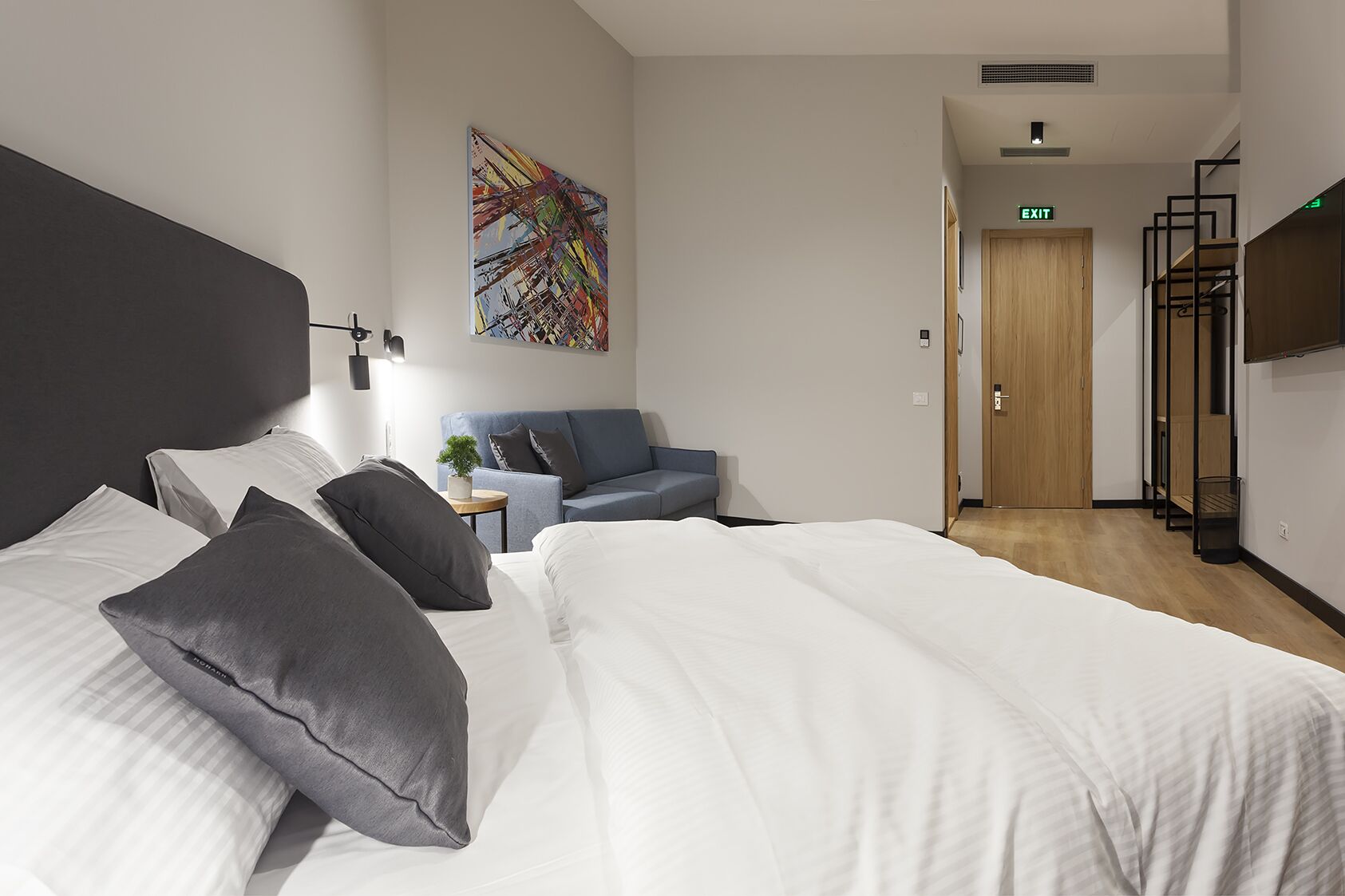 A modern hotel room with a large bed, a blue sofa, a small table with a plant, and a colorful abstract painting on the wall.