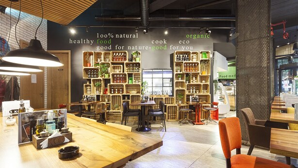 Interior de restaurante con mobiliario de madera, estantes llenos de botellas y un cartel que promociona alimentos naturales y orgánicos.
