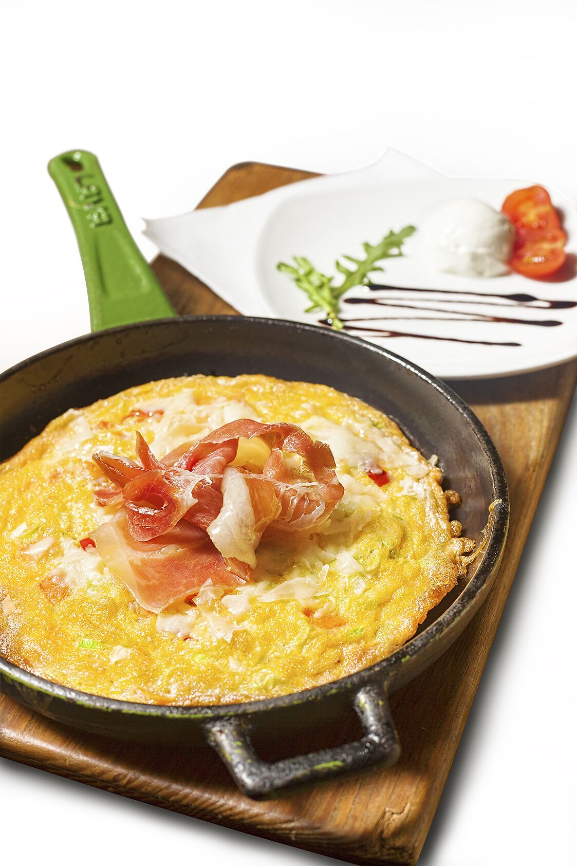 Een koekenpan met een gerecht, mogelijk een omelet, versierd met spek en tomaten, op een houten plank.